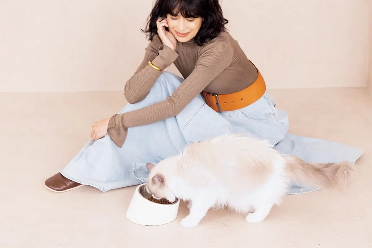 女性が、白い長毛猫の食事を見守っている。猫は白いボウルから食事をしている