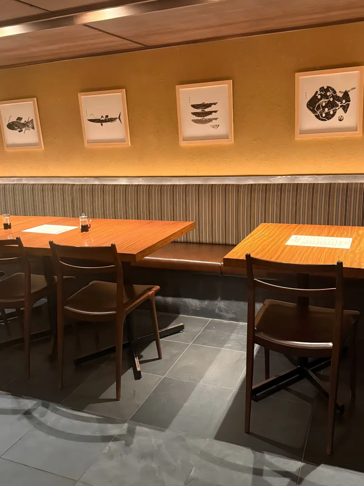 黄色の壁に魚の絵が飾られたレストランのテーブル席。木製のテーブルと椅子、灰色の床、背もたれ付きのベンチがある。
