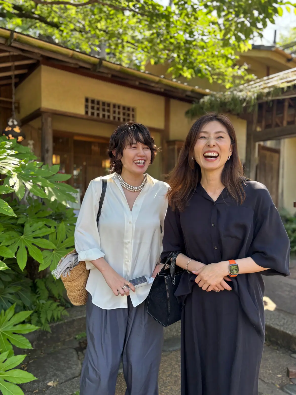 笑顔の女性2人が、伝統的な木造建築物の前で並んで立っている。左の女性は白シャツにグレーのパンツ、右の女性は黒いワンピースを着ている。緑豊かな植物が背景に見える。
