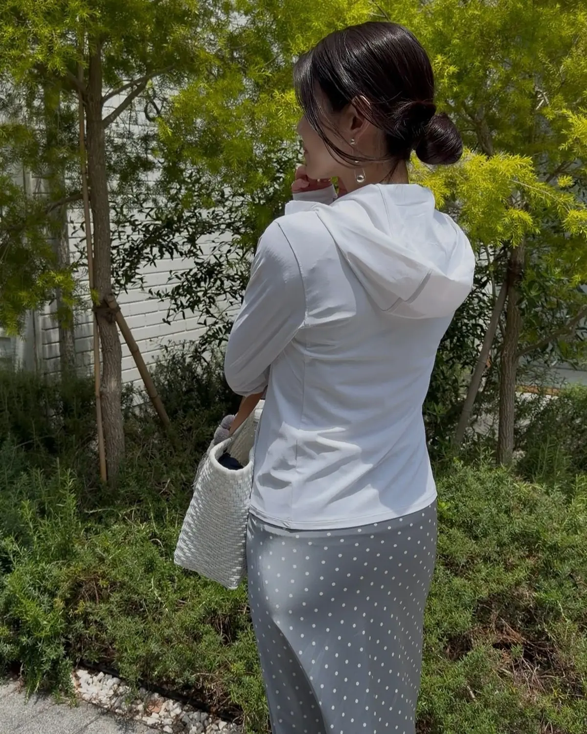 白のフード付きトップスとグレーに白ドット模様のスカートを着た女性が、屋外の庭園で背を向けた姿。左手にはグレーのトートバッグを下げ、耳にはイヤリングを着用。周囲には緑の低木やヤシの木が生い茂る。