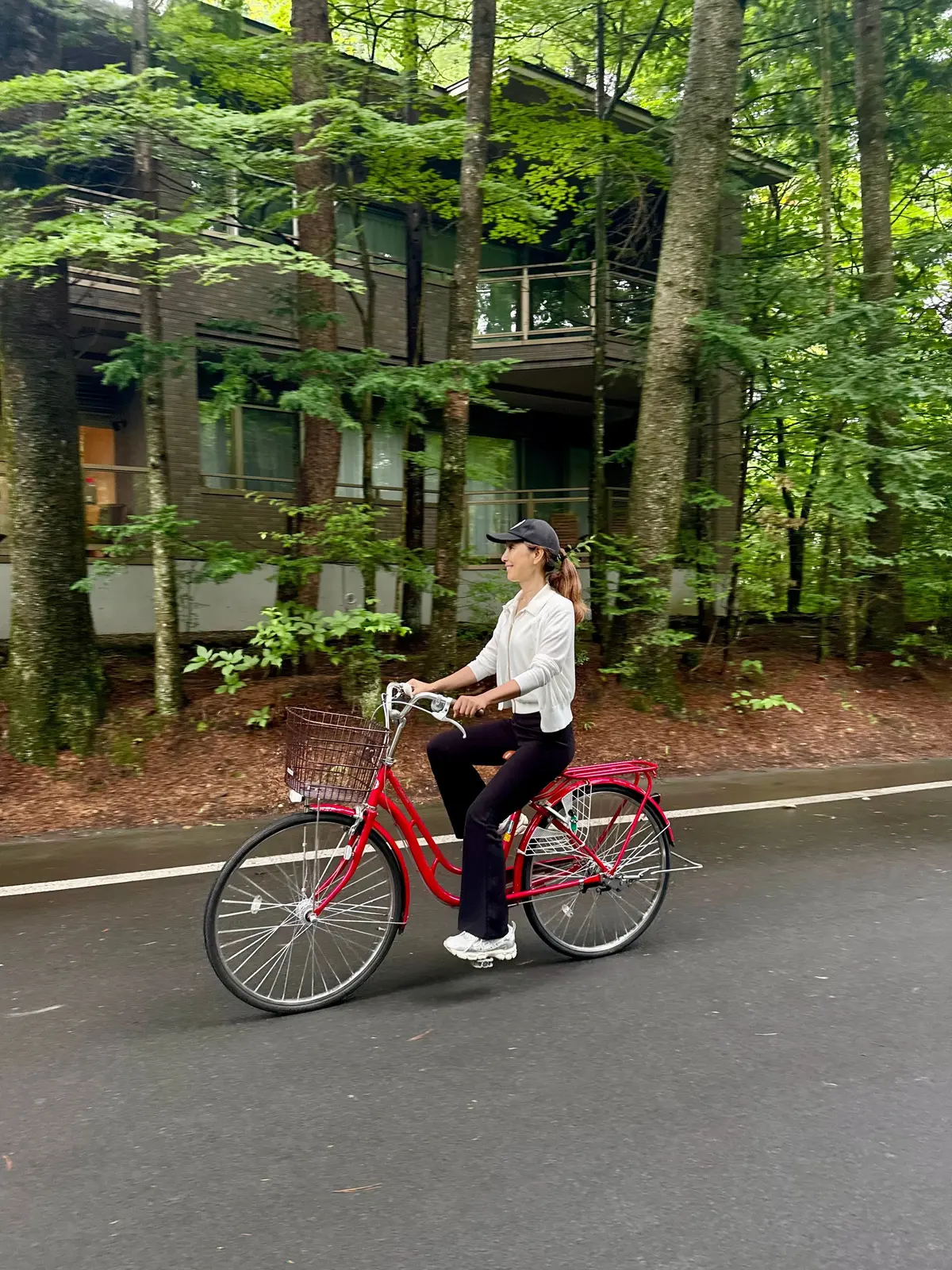 赤い自転車に乗った女性が、木々に囲まれた建物の前を走っている。女性は白いシャツに黒い帽子を着用し、黒いズボンとスニーカーを履いている。自転車には籐製のカゴが付いている。