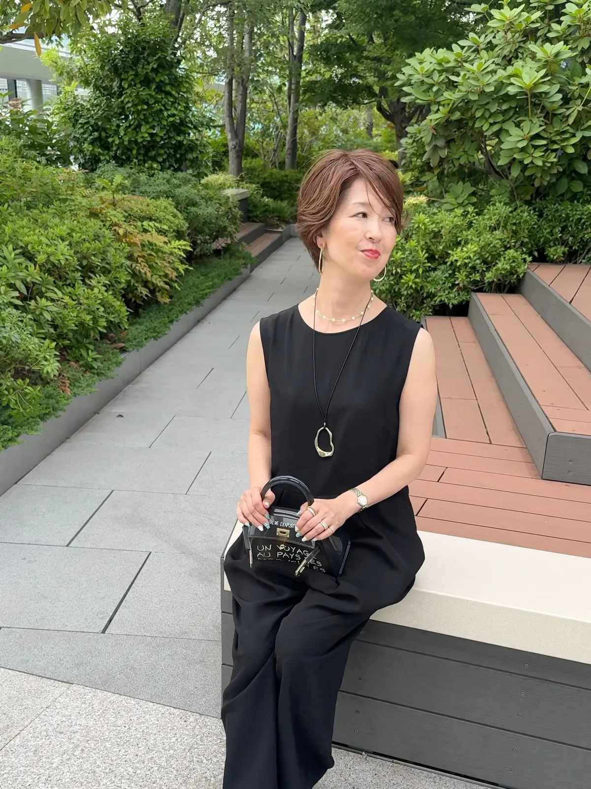 黒のワンピースで、屋外の庭園に座る女性。左手には黒のクラッチバッグ、右手には長いネックレスを身につけている。背後には緑の低木と木々が広がる。