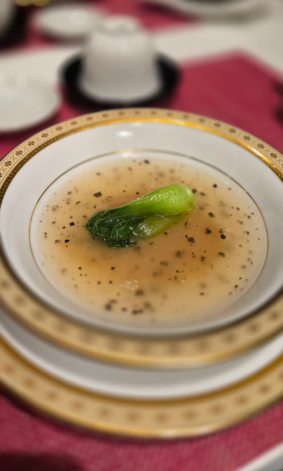 金縁の白い皿に、淡い茶色のスープが注がれており、その中央に緑色のボクチャイが置かれている。スープには黒い粒々が散らばっている。背景には、カップやソーサー、赤いテーブルクロスが見える。