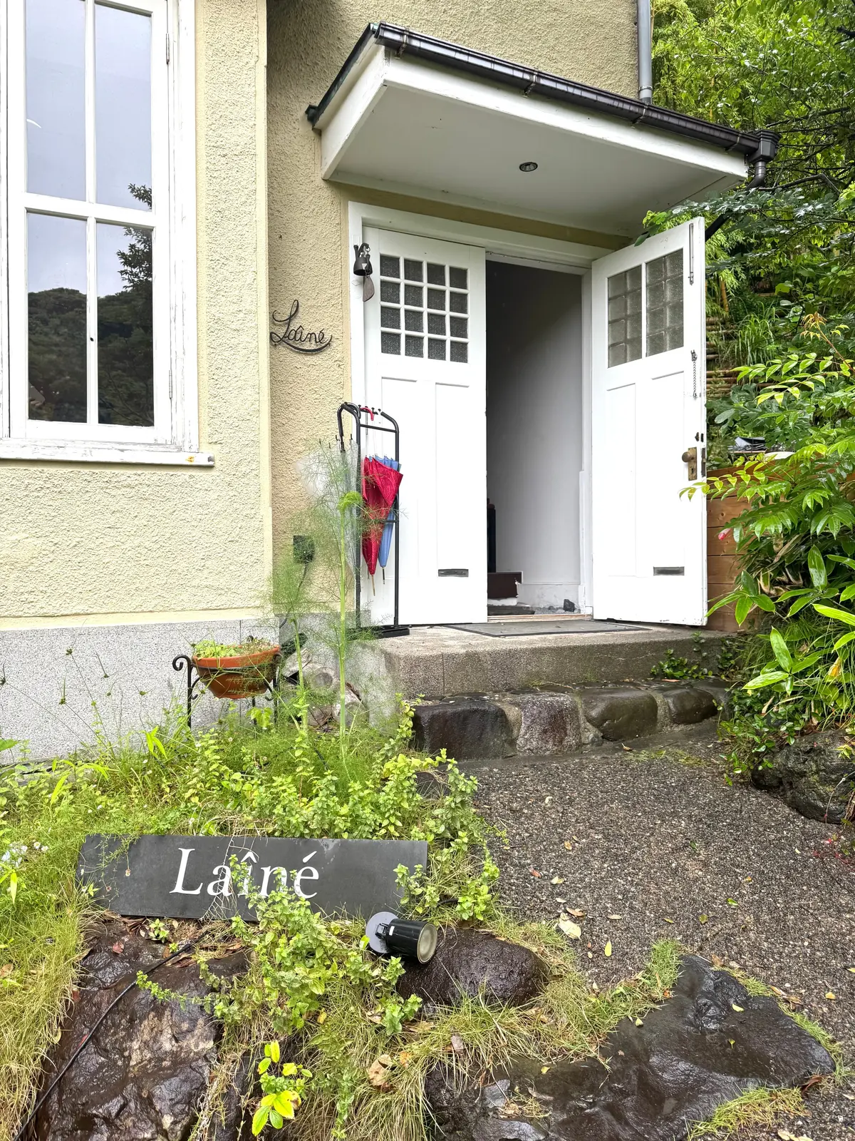 ベージュの家正面の入り口、石段を上がった場所に「Laîné」の看板があり、開いた白いドアから中が見える。手前に緑の植物と石が配置され、傘立てに赤と青の傘が掛かっている。