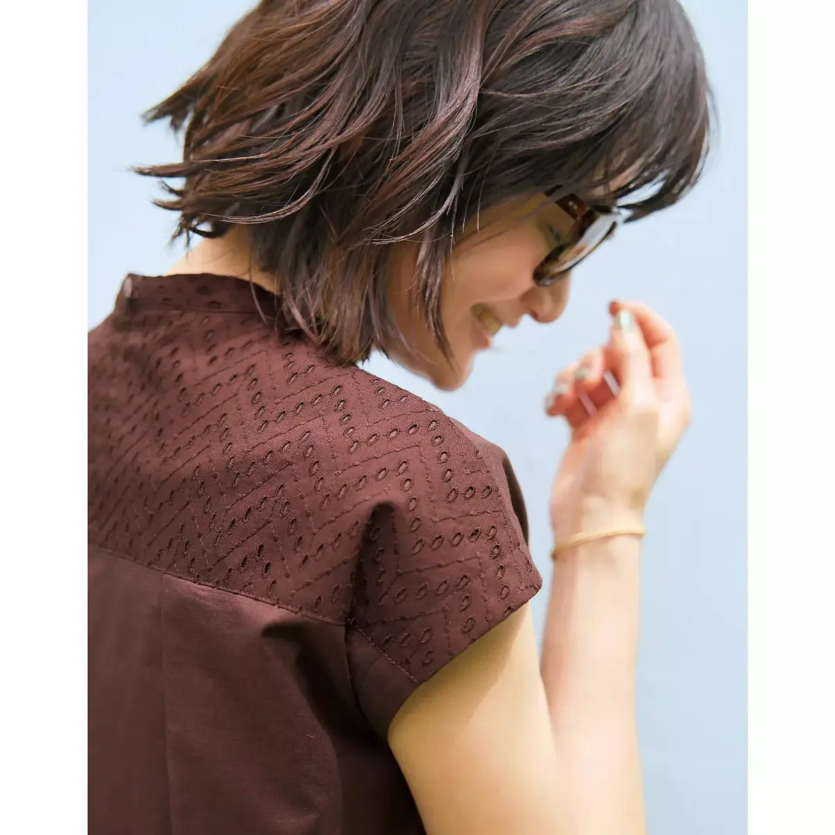 茶色のワンピースを着た女性が白い壁に寄りかかり、草むらに立っています。女性はサングラスをかけていて、右手に編みかごバッグを持っています。足元にはサンダルを履いています。