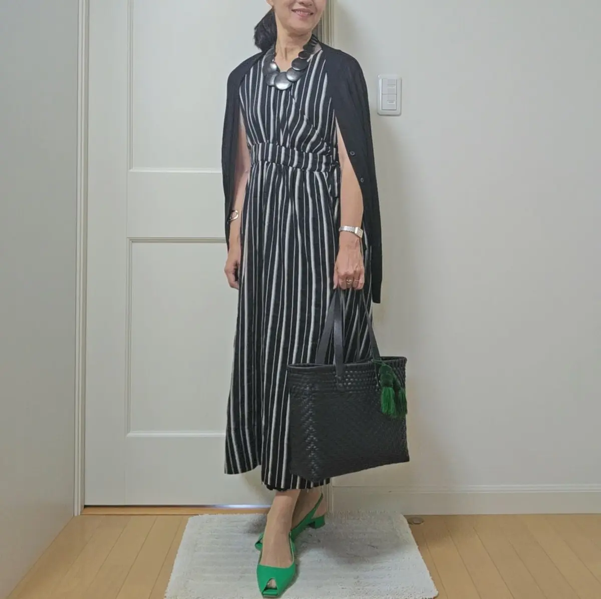 黒と白のストライプのミモレ丈ワンピースに、黒のカーディガンを羽織った女性。鮮やかな緑色のパンプスを履き、シルバーのネックレスと腕時計を身につけている。黒の編み込みバッグを提げ、白いドアの前に立っている。