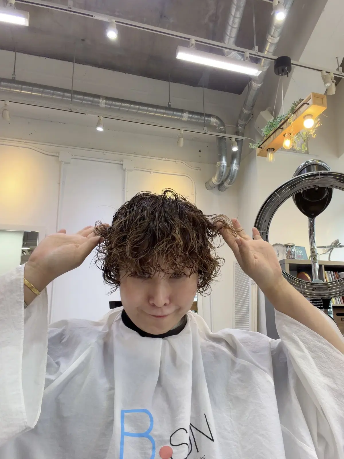 美容院でカットを受けた若い男女。肩までの長さのゆるいカールヘアで、白いカットクロスを着用。背景には大きな鏡と観葉植物が見える。
