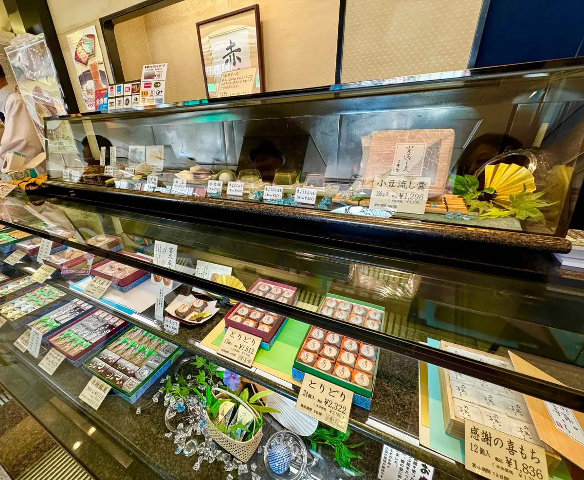 ガラスケースに並んだ様々なお菓子。抹茶色、赤、茶色など様々な色合いの和菓子が、大小様々な形で整然と並んでいる。上段には緑色の小分けになったお菓子、下段には円形や四角形の詰め合わせ菓子が並び、右下には「感謝の恵もももち」と書かれた商品がある。