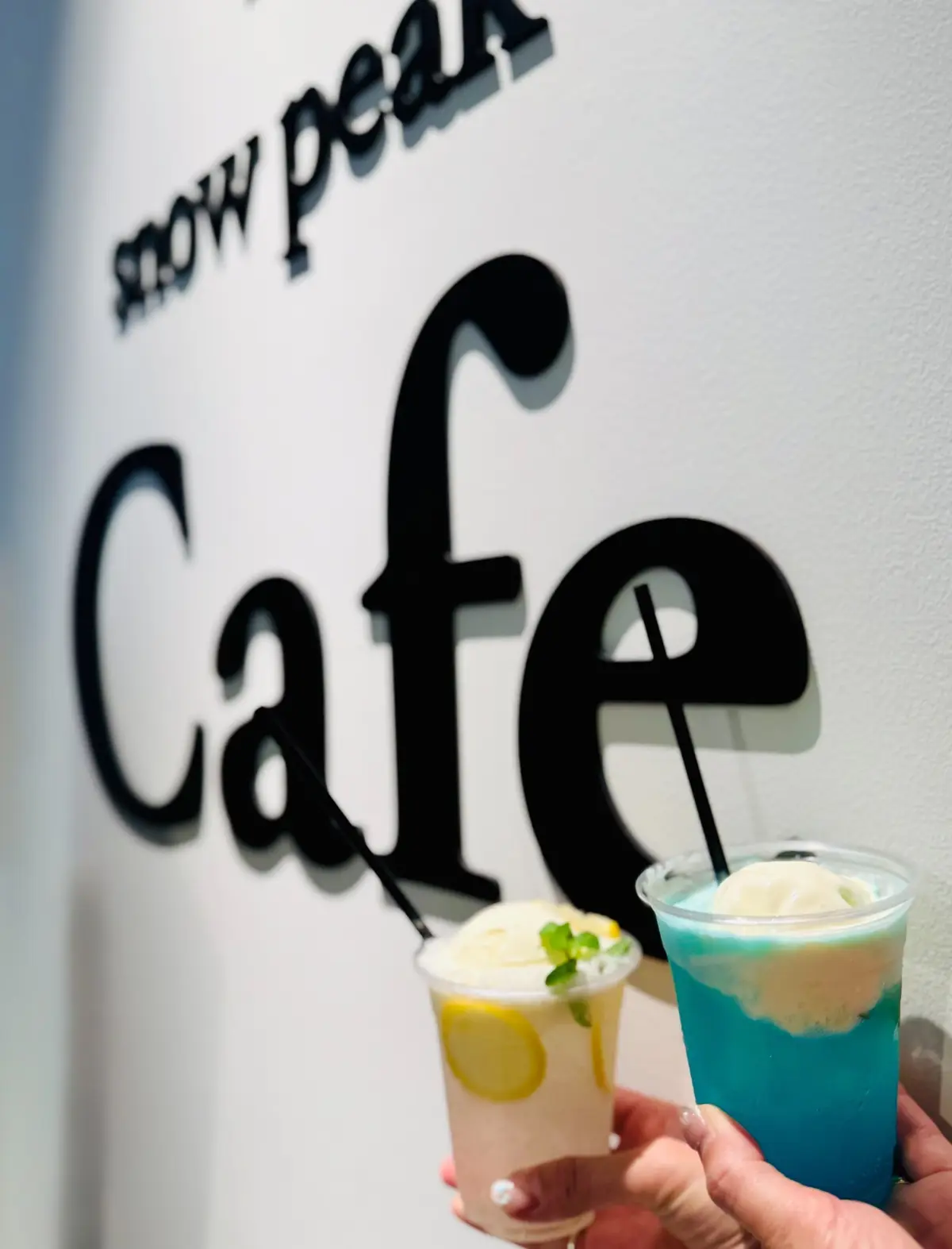 雪峰茶屋カフェの背景に、手で持たれた2つの透明なプラスチックカップに入ったドリンク。左はレモンとハーブが飾られた淡いピンク色のドリンク、右は鮮やかな青色のドリンク。両方とも白いクリームが乗っている。