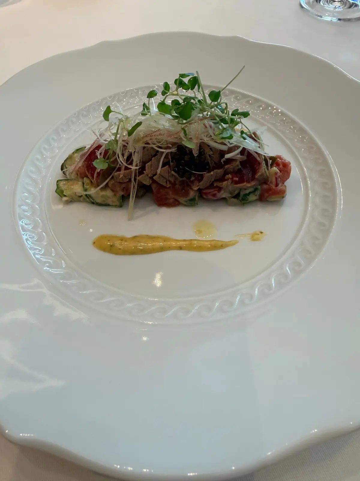 白磁の皿に盛り付けられた、赤身肉と野菜の盛り合わせ。肉は薄切りで、ズッキーニやハーブと共に円形に配置。横には黄色いソースが流れるように添えられ、上に微かな緑色の葉野菜が乗っている。
