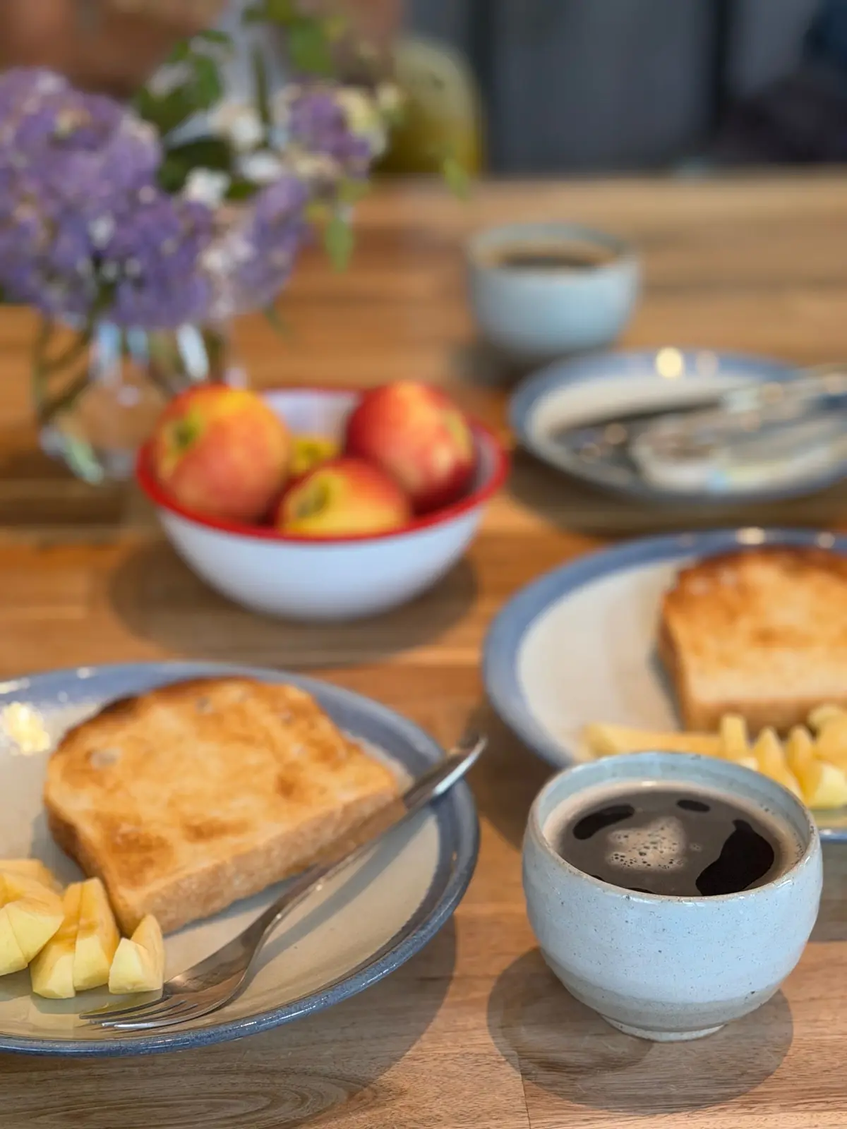 木製のテーブルに並べられた朝食。パン、フルーツ、コーヒー、花が配置されている。青色の縁取りがされた皿にパンとカットフルーツ、別の皿にはコーヒーが入った器が置かれている。赤と白のボウルには桃が並んでいる。
