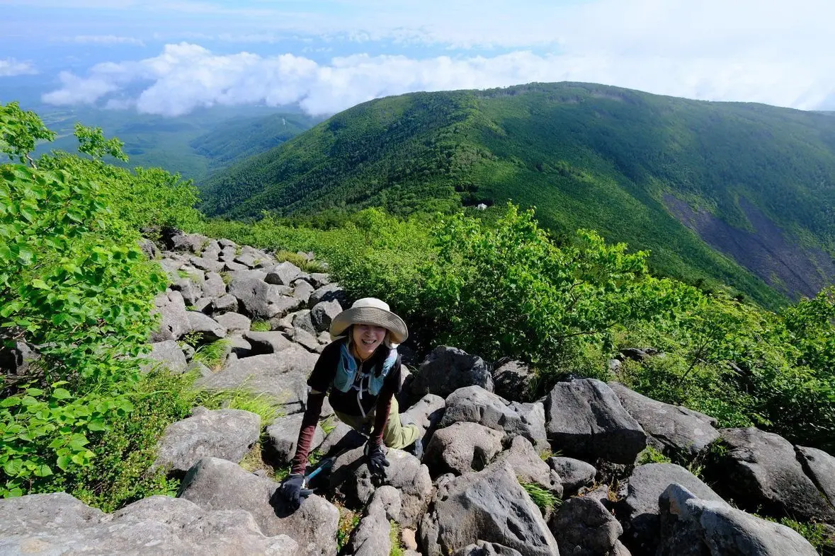 緑豊かな山々と雲海を背景に、帽子をかぶった女性が岩場を登っている様子。笑顔でカメラ目線。