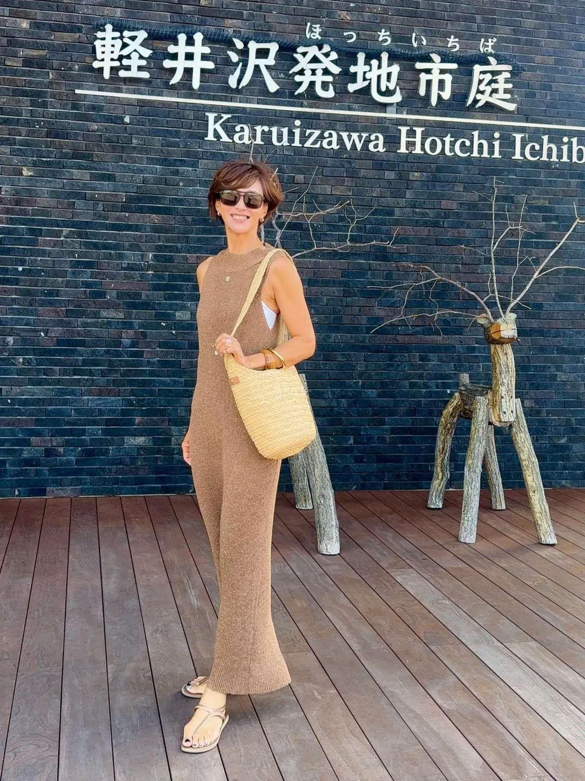 木製のオブジェを前にポーズをとる女性。背景には「Karuizawa Hotchi Ichiiba」と書かれた黒いレンガの建物。茶色のワンピースを着て、ベージュのバッグと靴を身につけている。