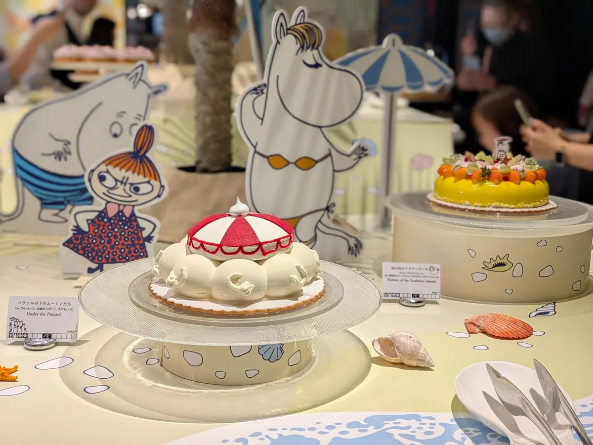 ムーミンキャラクターが描かれた紙製の立体ディスプレイと、赤と白の傘が乗ったムースケーキ、黄色いフルーツケーキが並んだテーブル