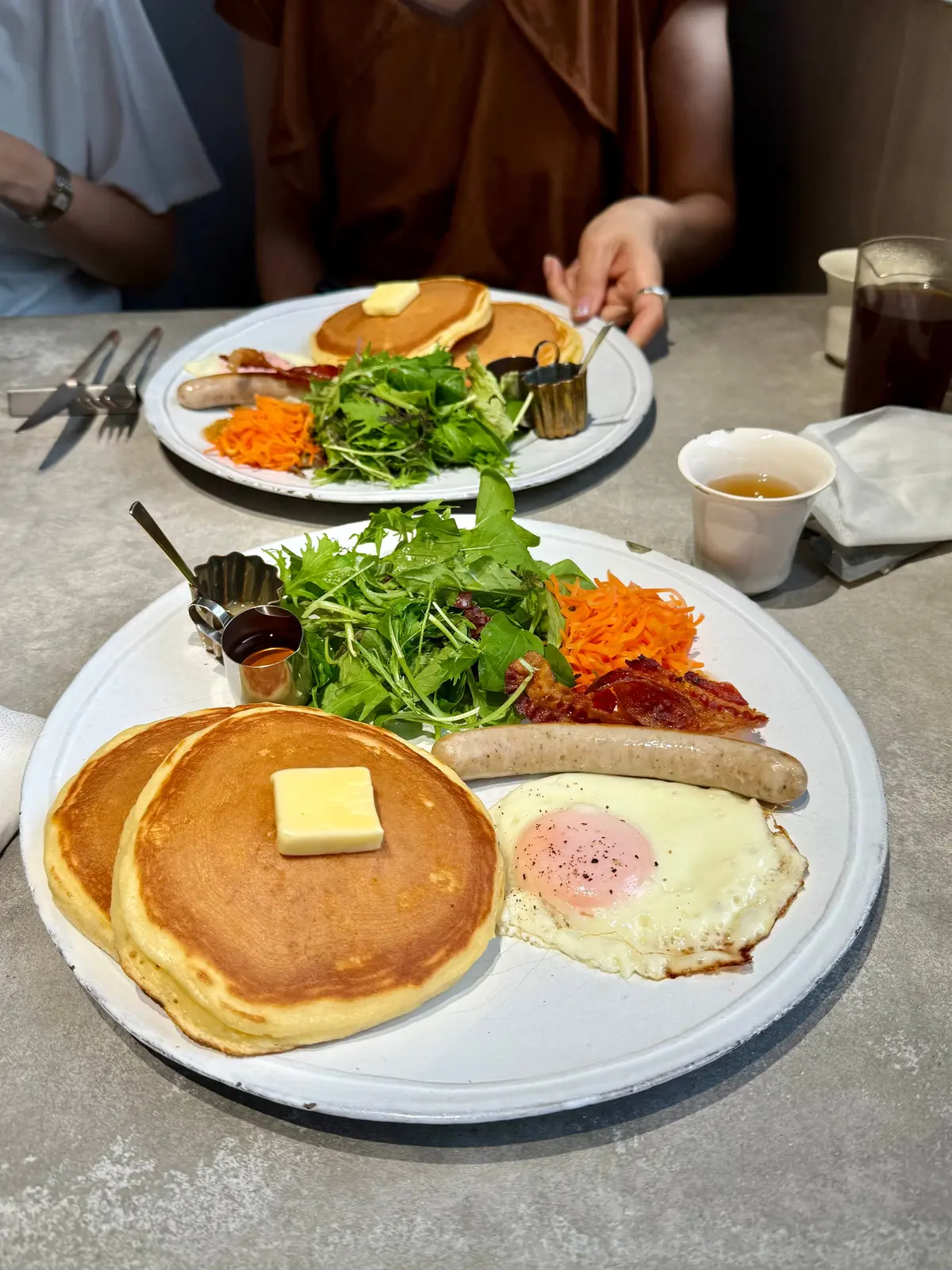 グレーのテーブルに置かれた、パンケーキ、ソーセージ、目玉焼き、サラダの朝食プレート。パンケーキにはバターが乗っており、サラダにはドレッシングが添えられている。隣には飲み物と小さなカップが置かれている。