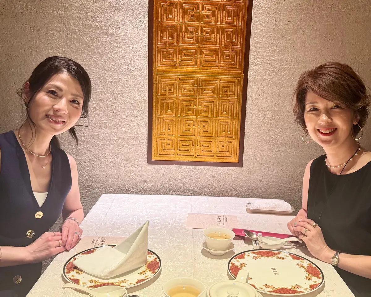 二人の女性がテーブルに座り、茶碗と料理が置かれたテーブルクロス付きのテーブルで笑顔で会話している。背景には木製の装飾パネルがある。