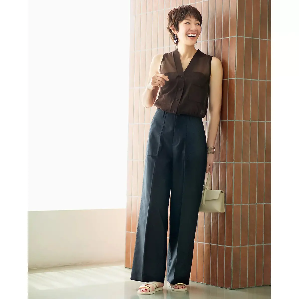 茶色のボタン付きノースリーブトップスに、濃い紺色のワイドパンツを合わせた女性。右手にベージュのハンドバッグを持ち、笑顔でこちらを見ている。背景はテラコッタ色のタイル壁。足元はベージュのサンダル。