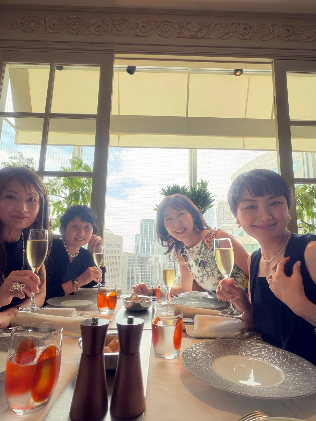 窓から都市の景色を望む、4人の女性がテーブルを囲んで食事を楽しんでいる様子。シャンパンやジュース、料理が並び、笑顔で談笑している。