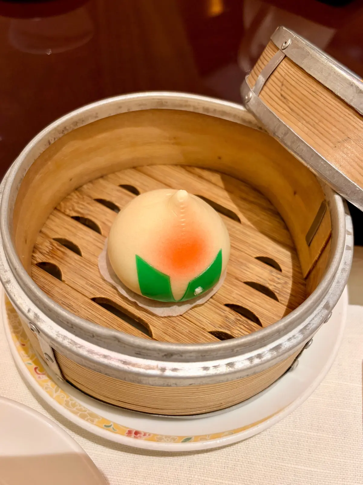 木製の蒸し器に、緑と赤の模様が付いた白い蒸しパンが1つ置かれている