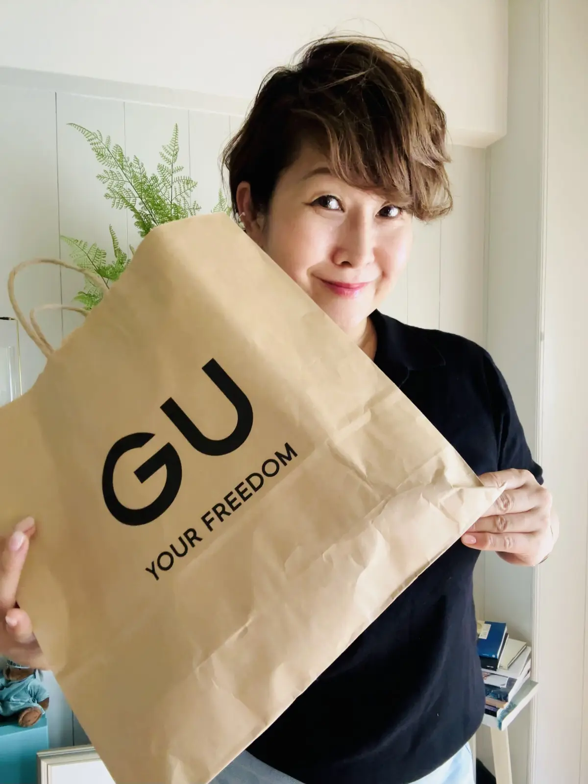 女性が紙袋を手に持つ。袋には「GU」と「YOUR FREEDOM」という文字が印刷されている。背景には観葉植物と棚があり、明るい室内に置かれている。