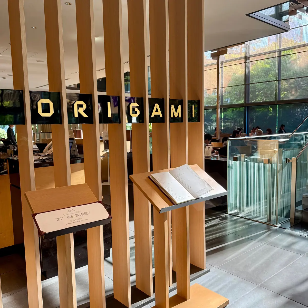 キャピトルホテル東京といえばオリガミ。木製の垂直スラットが並び、その間に黒い文字で「ORIGAMI」と書かれたサインがある。スラットの前にメニュー立てが置かれ、メニューが挟まれている。背景には窓から外の緑が見える。