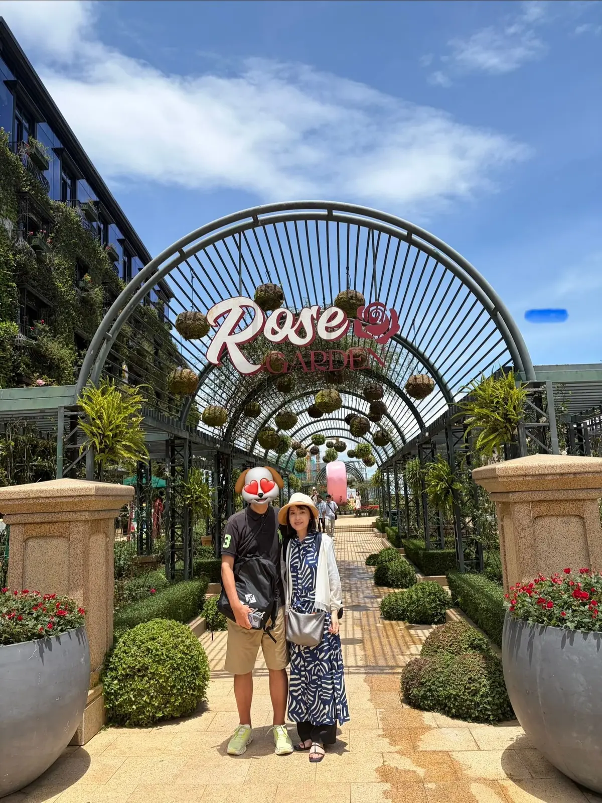 ベトナム・ダナンの人気観光地「フランス村」にあるローズガーデン前の2人。アーチ型のゲートには「Rose Garden」の文字。左右に植栽があり、青空の下、白い雲が浮かんでいる。