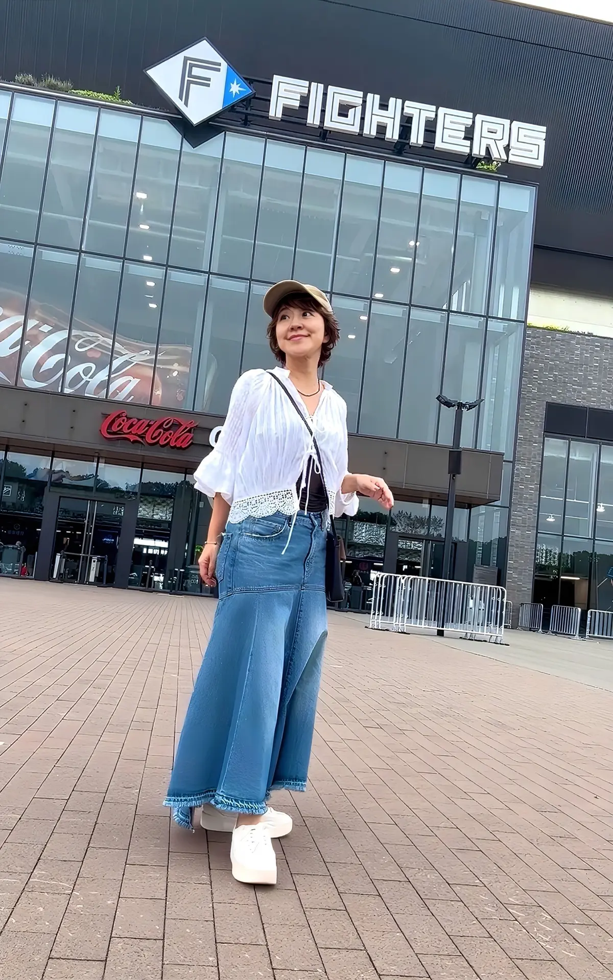 白いシャツにデニムのスカート、キャップをかぶった女性が、正面に Fighters のロゴが入った建物の前でポーズをとっている。背景には Coca-Cola のロゴも見られる。