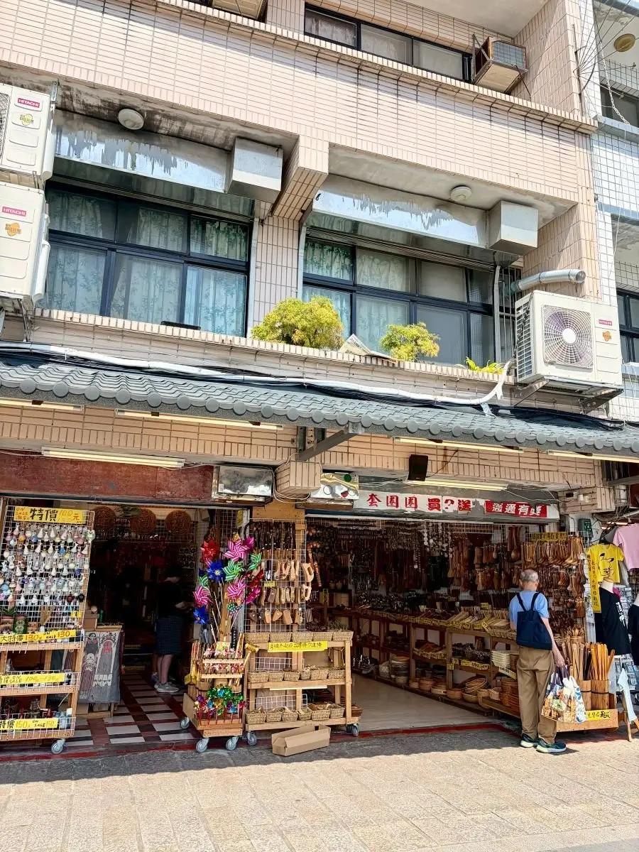 伝統的な建物前に広がる手工品マーケットで、木製の小物や編み物などが並び、顧客が商品を眺めている。