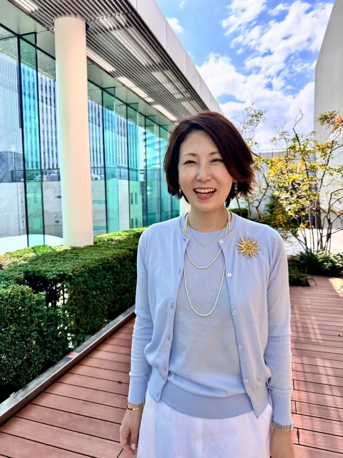 女性が明るい笑顔で屋外のテラスに立っている。淡い青のセーターに真珠のネックレス、金のブローチを身につけ、背景にはガラス張りの建物と手入れのされた低木が広がる。