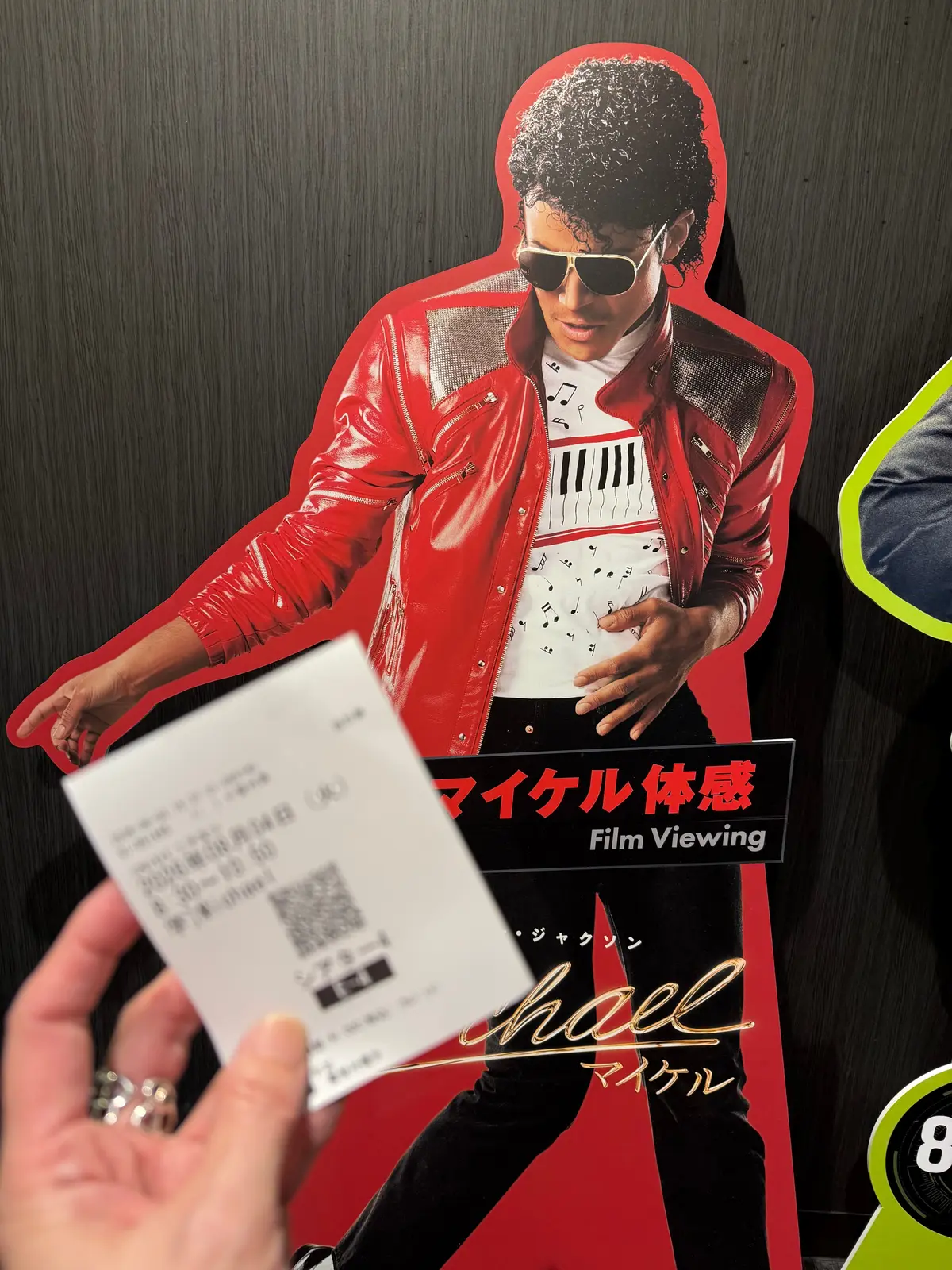 マイケル・ジャクソンの立像ポスターとチケット。赤色のジャケットを着用したジャクソンが右手を上げてポーズをとっている。ポスター下部には「Film Viewing」の文字と、マイケル・ジャクソンの名前が書かれている。手にはチケットが握られている。