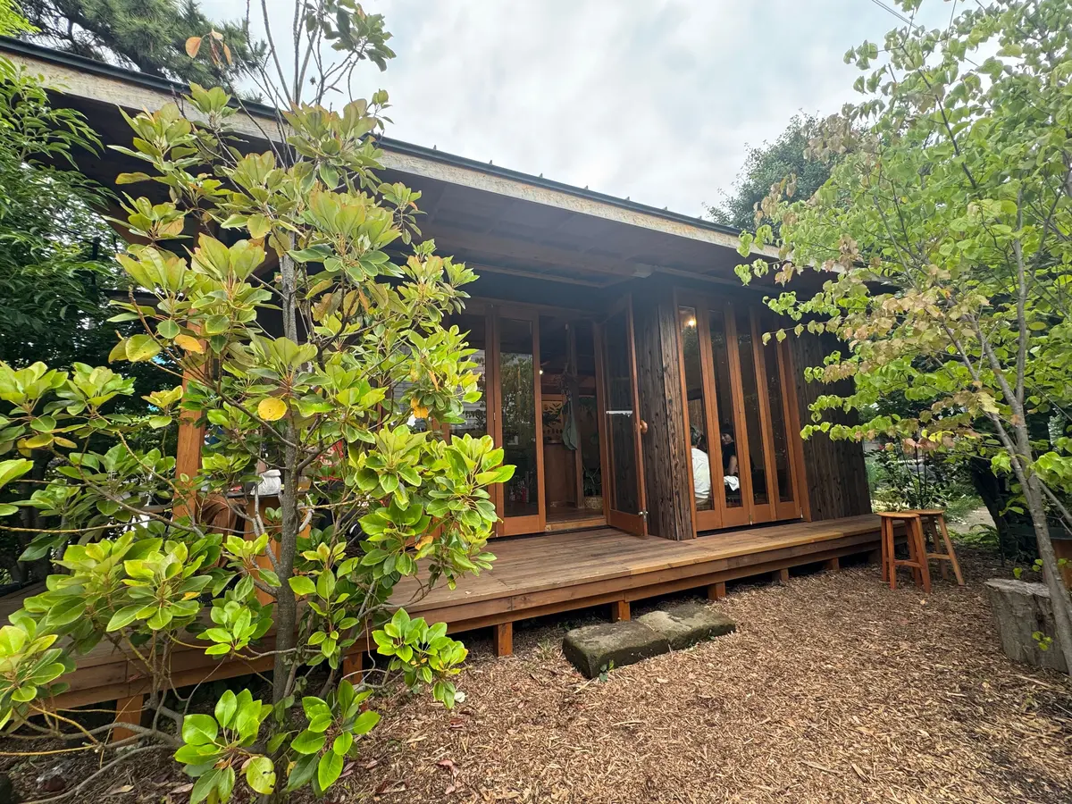木製の簡易小屋が木製のデッキに設置され、周囲は木の葉が茂る庭園。ガラスのドアが開いて中が見え、中に人がいる。デッキには木製の椅子とテーブルが置かれている。