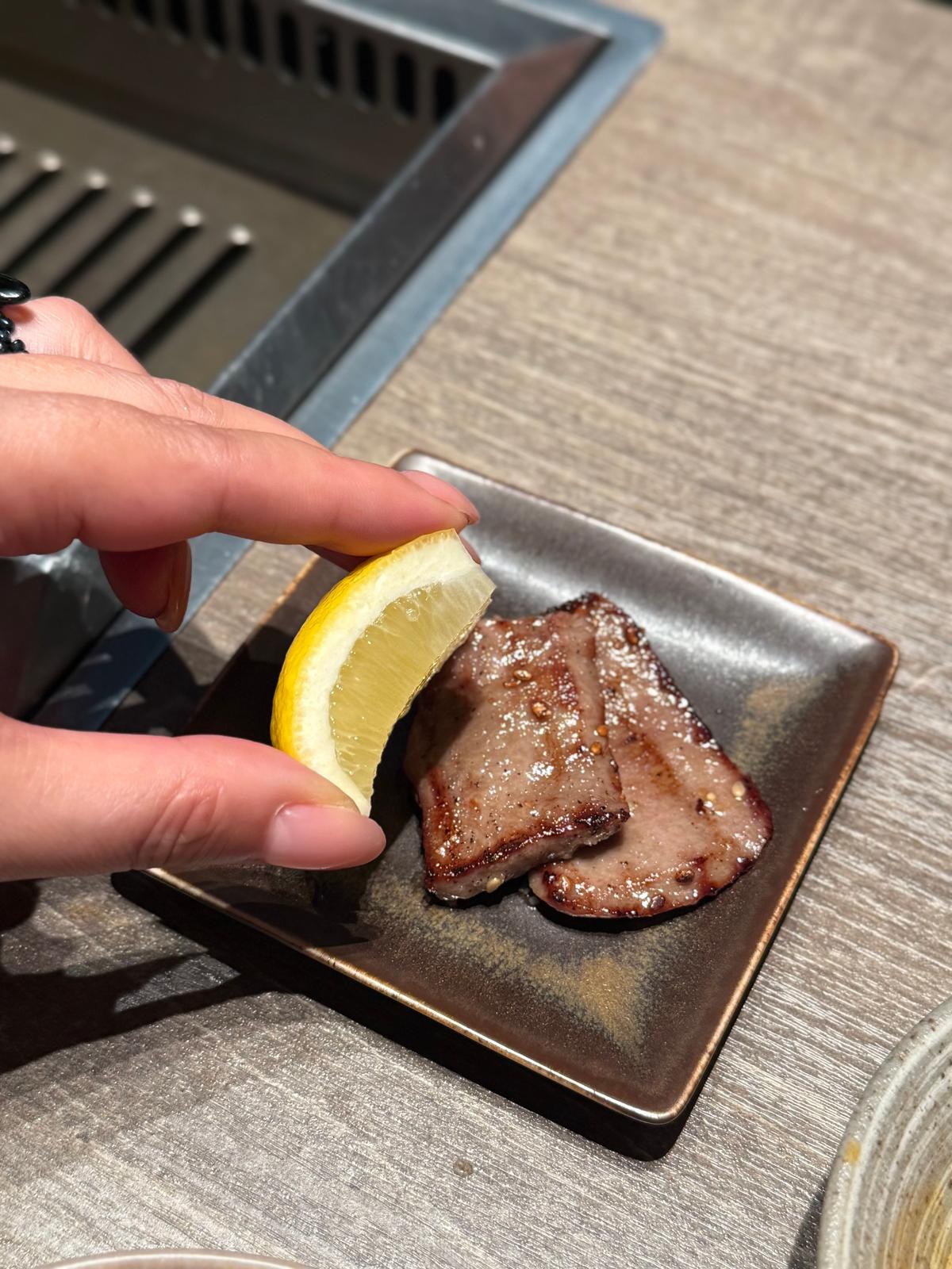 焼けた牛肉とレモンのウェッジが、茶色の正方形の皿に置かれている。手はレモンに触れている。背景には金属製のグリルがある。
