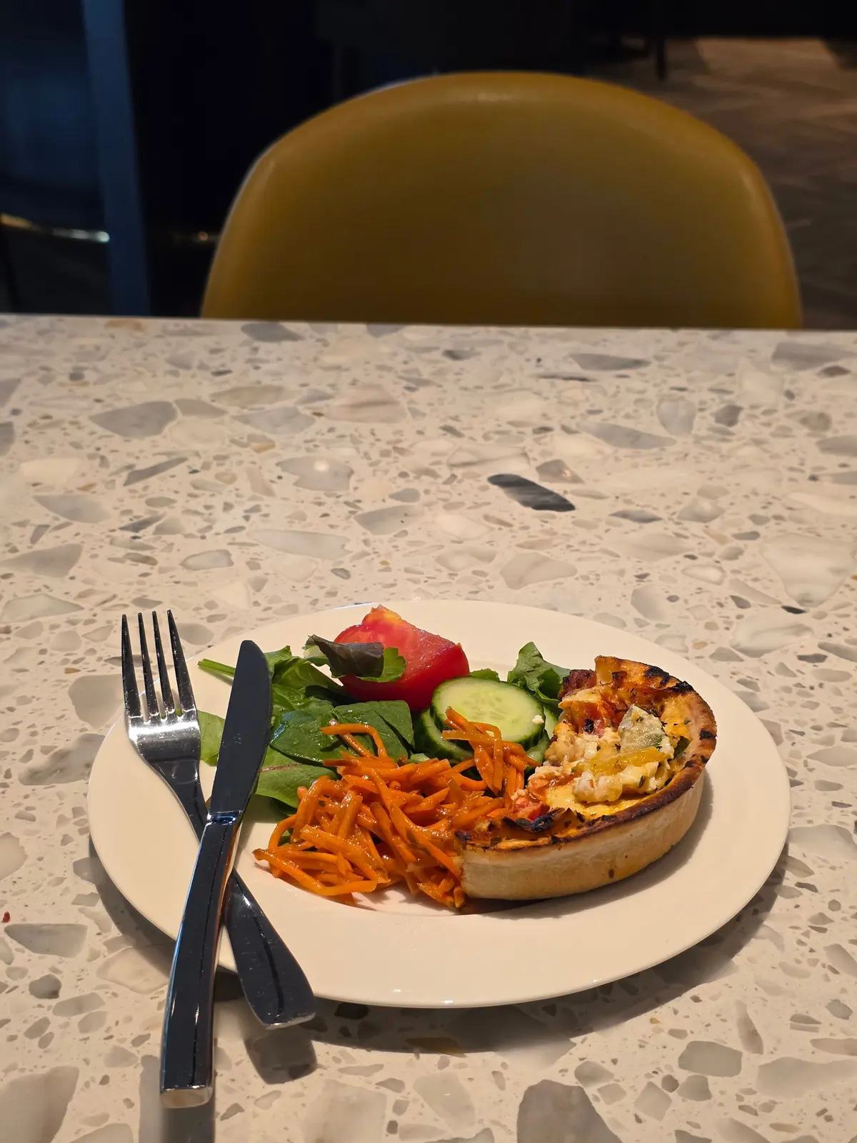 白い皿に、キッシュ、サラダ、カット野菜が盛り付けられている。背景には黄色の椅子と、テラゾー模様のテーブルがある。フォークとナイフが置かれている。