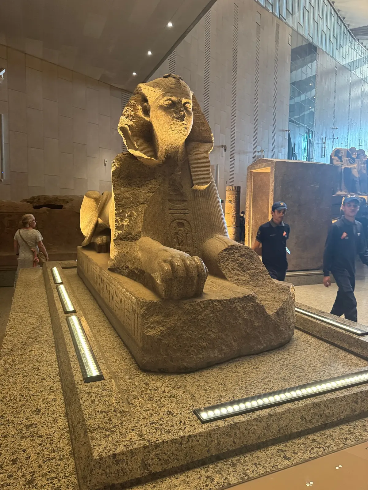 エジプトの博物館に展示された、ライオンの胴体に人間の顔を持つ巨大な石造りのsphinx像。正面を向いており、台座に置かれている。背景には人々や他の展示物が見える。