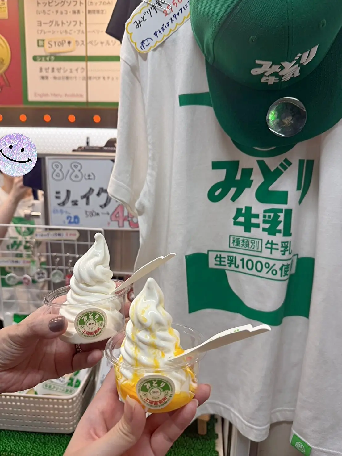 緑色のTシャツを着た手が、メリー乳業のソフトクリームを持っている。ソフトクリームは透明なカップに入っており、底にはオレンジ色のソースが塗られている。背景にはメリー乳業の広告とメニューが見える。