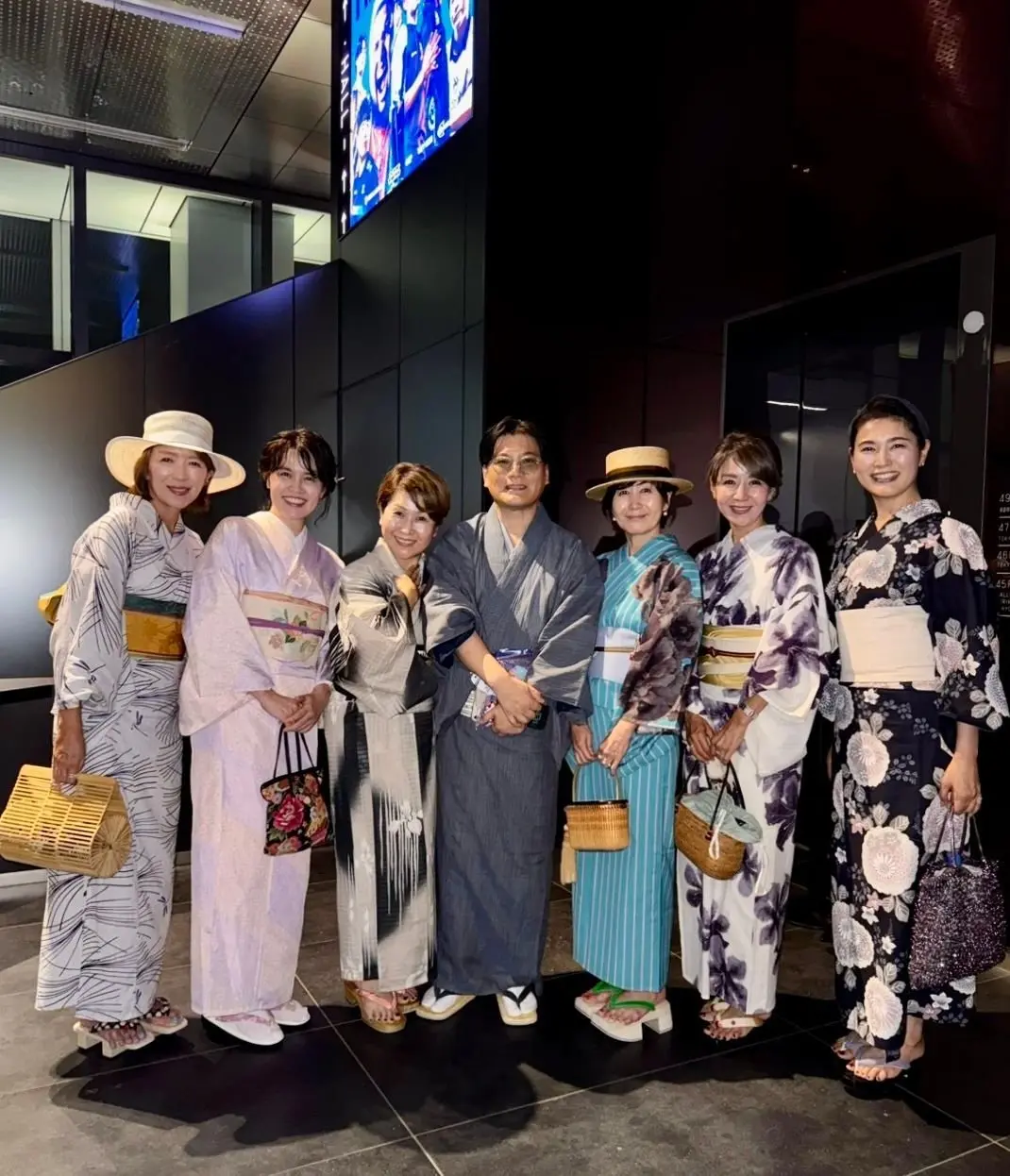6人の男女が、青や白、紺などの色合いの着物と浴衣を着て立っている。女性は麦わら帽子やバッグ、男性は盆履を着用。背景には黒色の壁と、青色のデジタルアートが映っている。