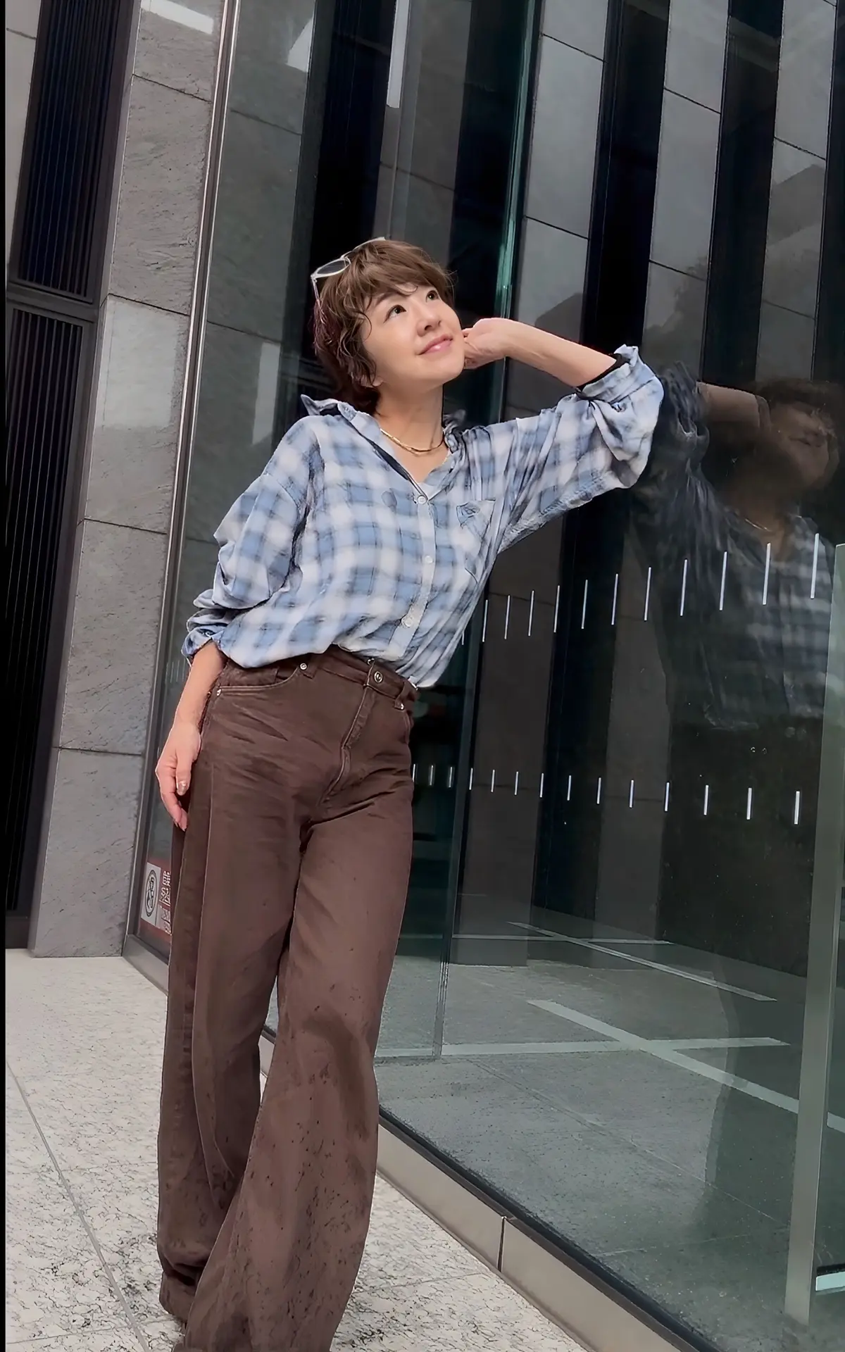 
茶色のワイドパンツにチェック柄のシャツを着た女性。右手を頭に当て、窓に映る自分の姿を見ている。背景は現代的な高層ビルの壁。
