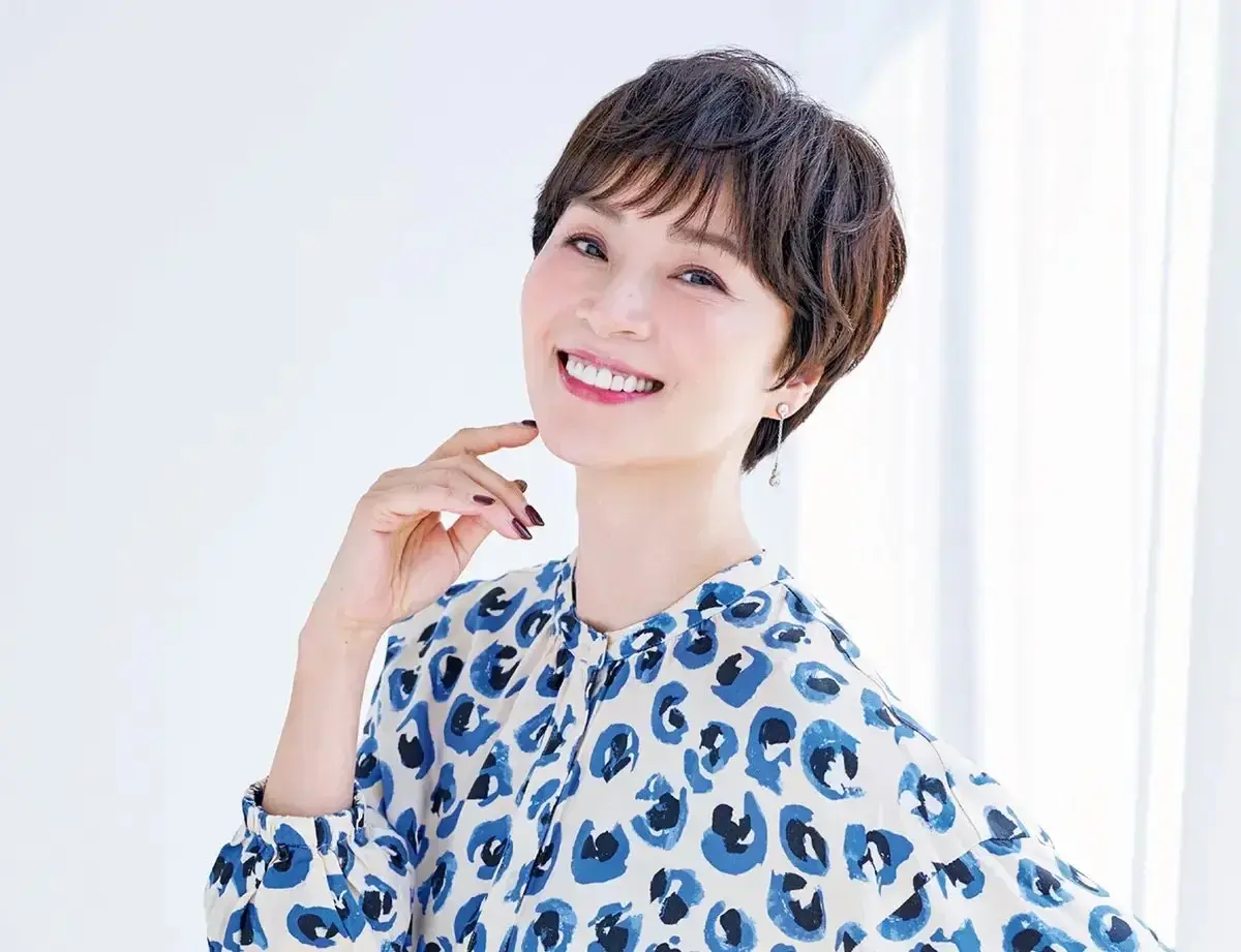 白い背景に、ショートカットの女性が青と白のヒョウ柄のブラウスを着て、ほほをくすぐるような表情で微笑んでいる。