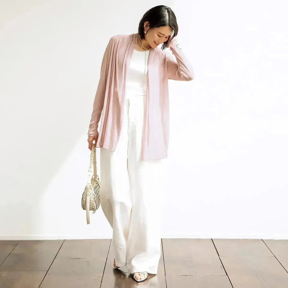 ピンクのロングカーディガンに白のワンピースを着た女性が、左手で髪を整え、右手には編みかごのショルダーバッグを下げています。