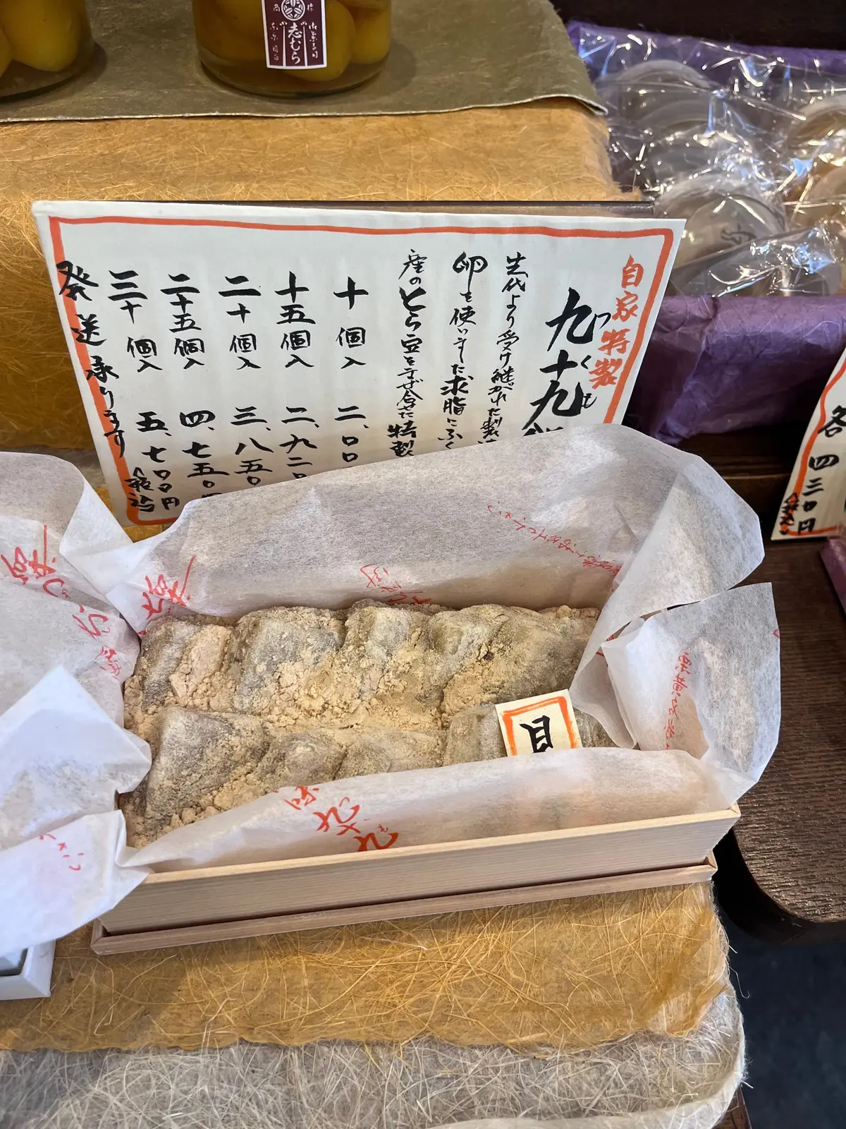 木製のトレーに、白菜漬け、梅干し、紫色の野菜、価格表が置かれている様子。白菜漬けは紙で包まれ、梅干しは瓶に入っている。価格表には日本語で商品名と値段が記載されている。