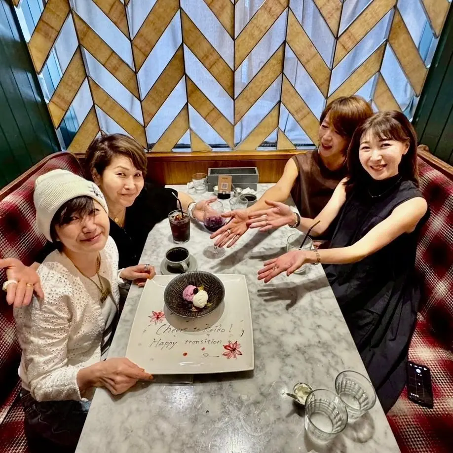 3人の女性がカウンター席に座り、ケーキと飲み物を伴うお祝いテーブルで笑い合う様子。中央のプレートには「Cheers to Reiko! Happy transition」と書かれている。