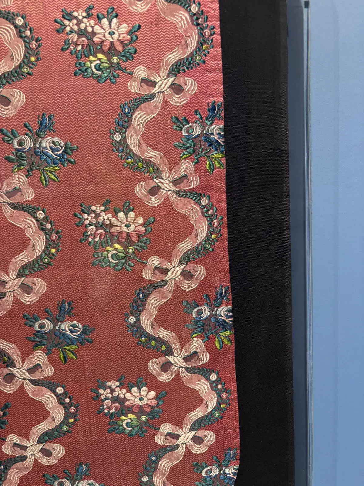 赤茶色の地に、花や葉、リボンが刺繍された模様が繰り返し描かれている。リボンはねじれながら配置され、全体的に華やかな印象。