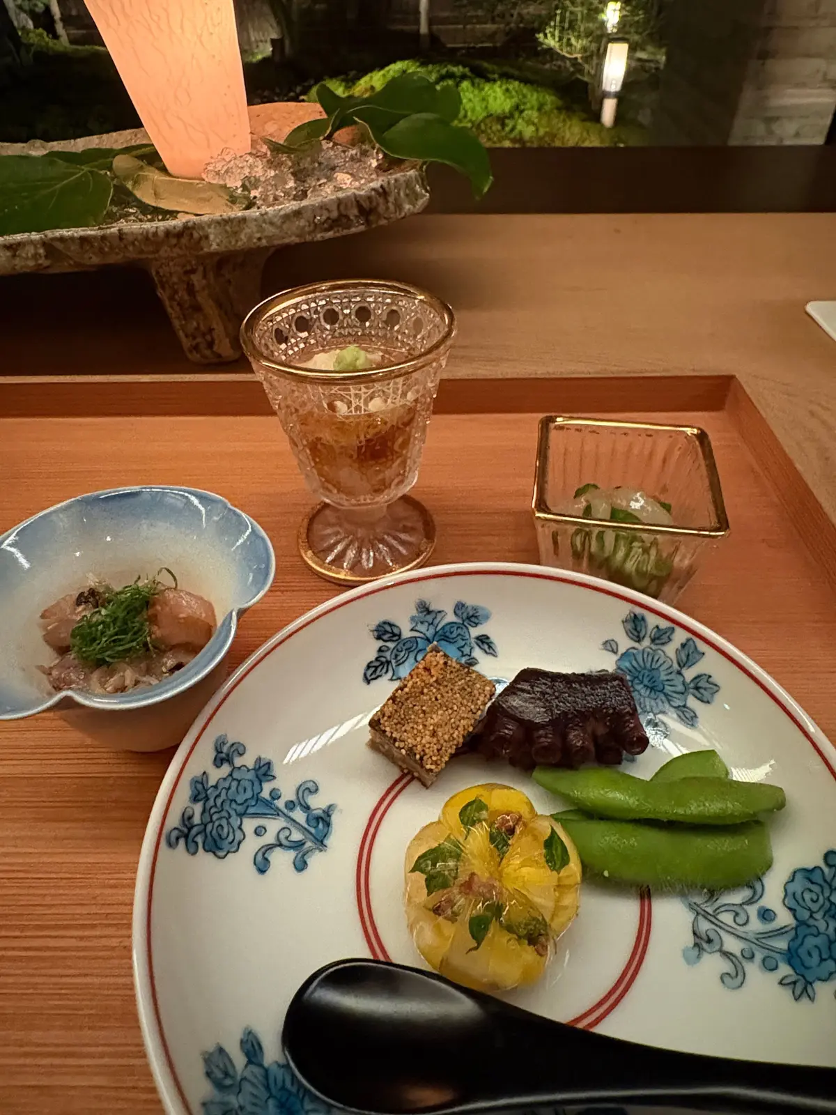 木製のテーブルに置かれた、和食のコース料理。白地に青い模様の皿には、様々な食材が盛り付けられている。小皿には魚料理、グラスにはお酒、小さな箱にはお新香らしきもの。背景には植物とキャンドル。