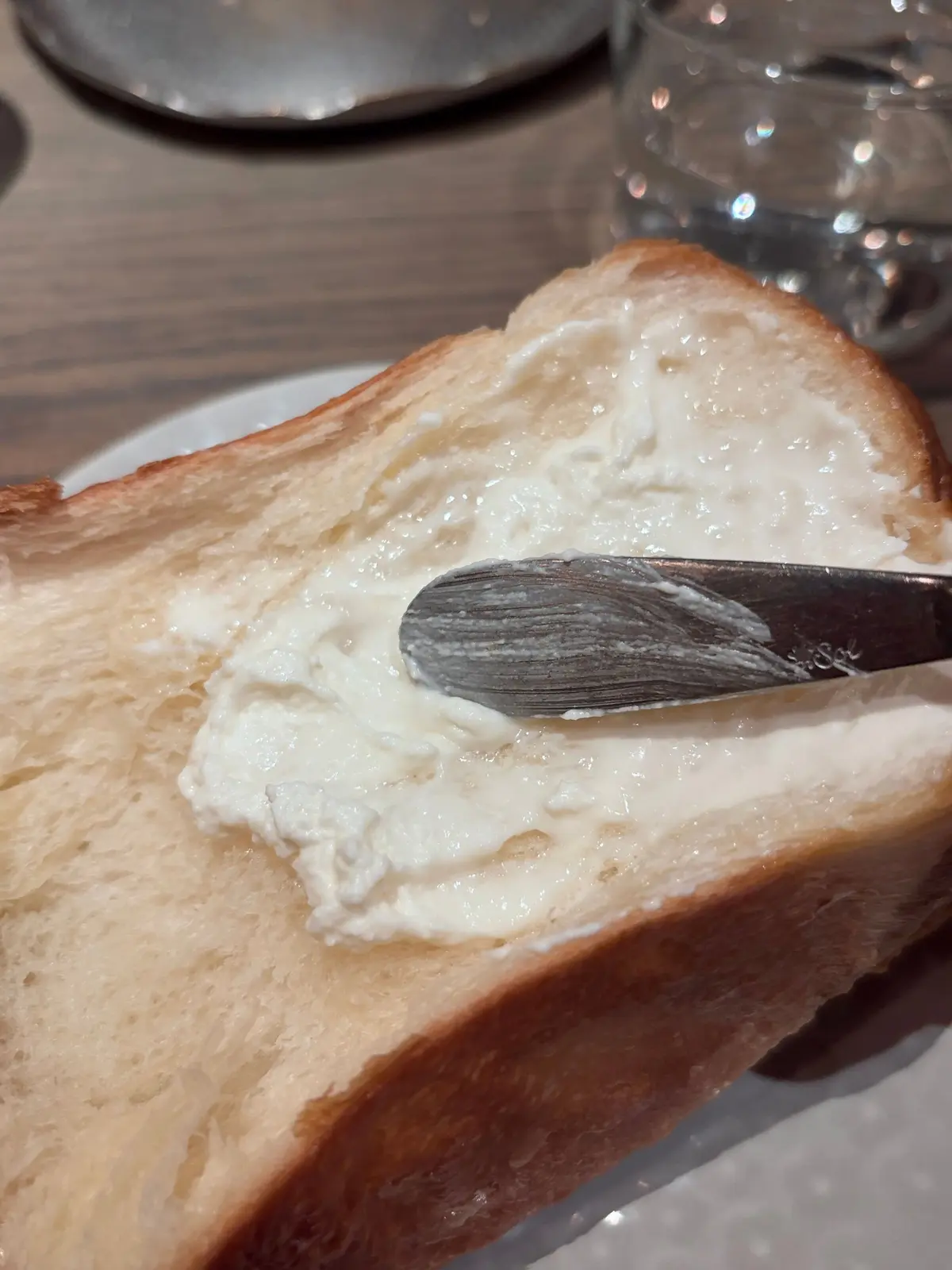 焼いた食パンにバターが塗られている様子。バターナイフが塗られたばかりのバターの上に置かれている。背景には水が張られたグラスと食器が見える。