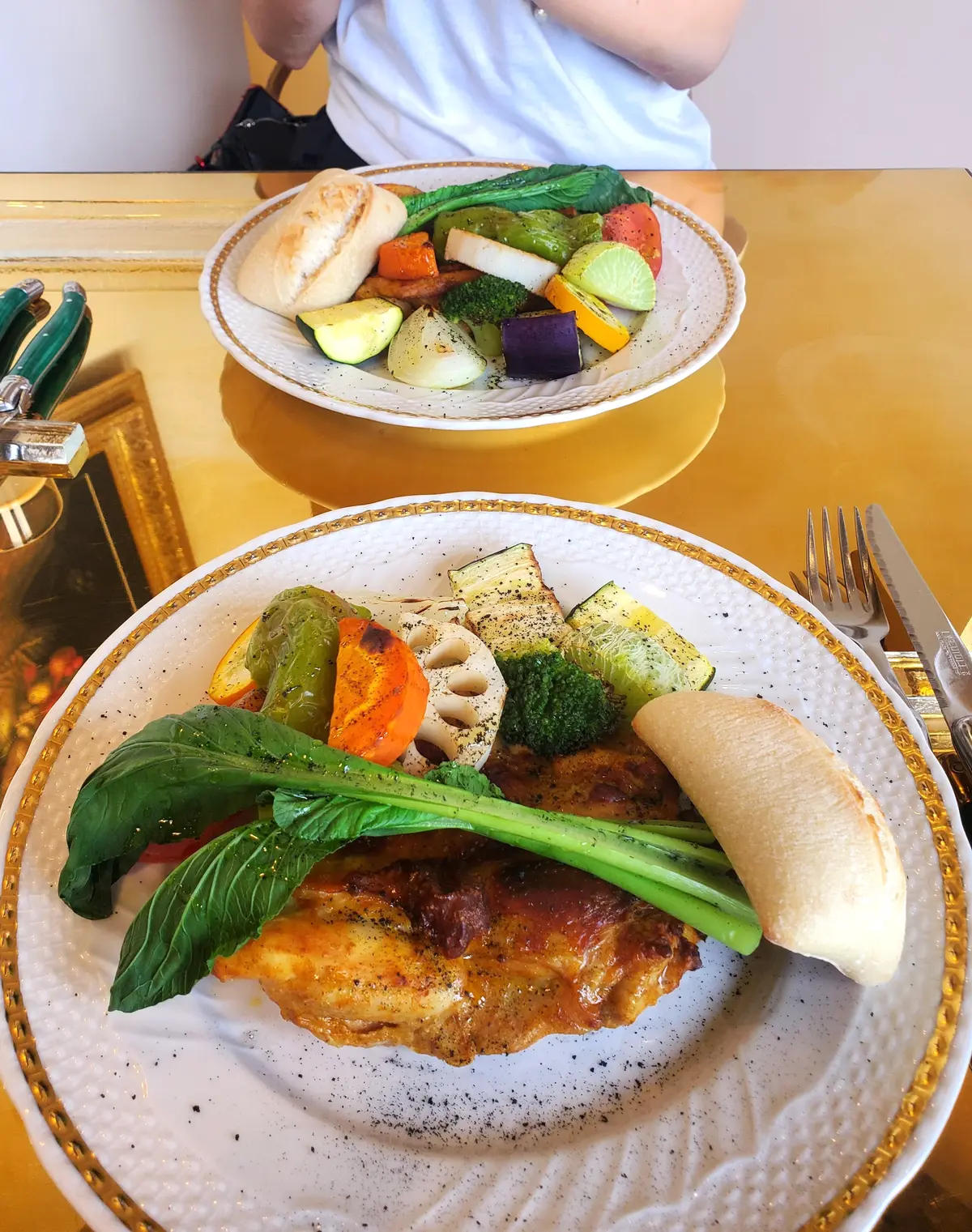 金色のテーブルに置かれた2つの白いお皿。それぞれに、鶏肉料理と彩り豊かな焼き野菜、パンが盛り付けられている。