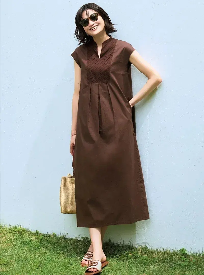 茶色のワンピースを着た女性が、青い壁のそばに立ってポーズをとっている。ワンピースは膝下丈で、胸元にレースがあしらわれている。女性はサングラスをかけ、手に麦わらのバッグを持っている。背景には緑の芝生が広がっている。