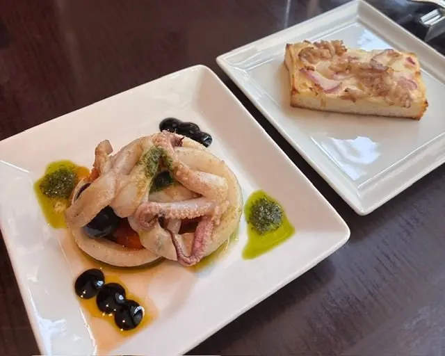白磁の正方形皿に盛り付けられたイタリア料理。左皿には揚げたイカの輪と、黒オリーブ、緑色のソース、バジルペーストが配置されている。右皿には、玉ねぎとチーズが乗った四角いパンが置かれている。