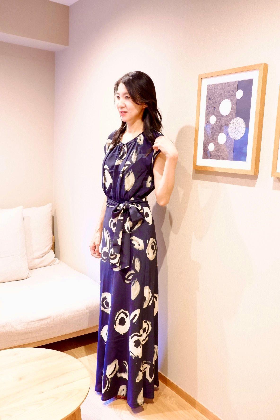 紺色の花柄ロングドレスを着た女性が、ベージュのソファの横で立っている。右壁には額縁に入った抽象画が飾られ、床には丸い木製のテーブルがある。