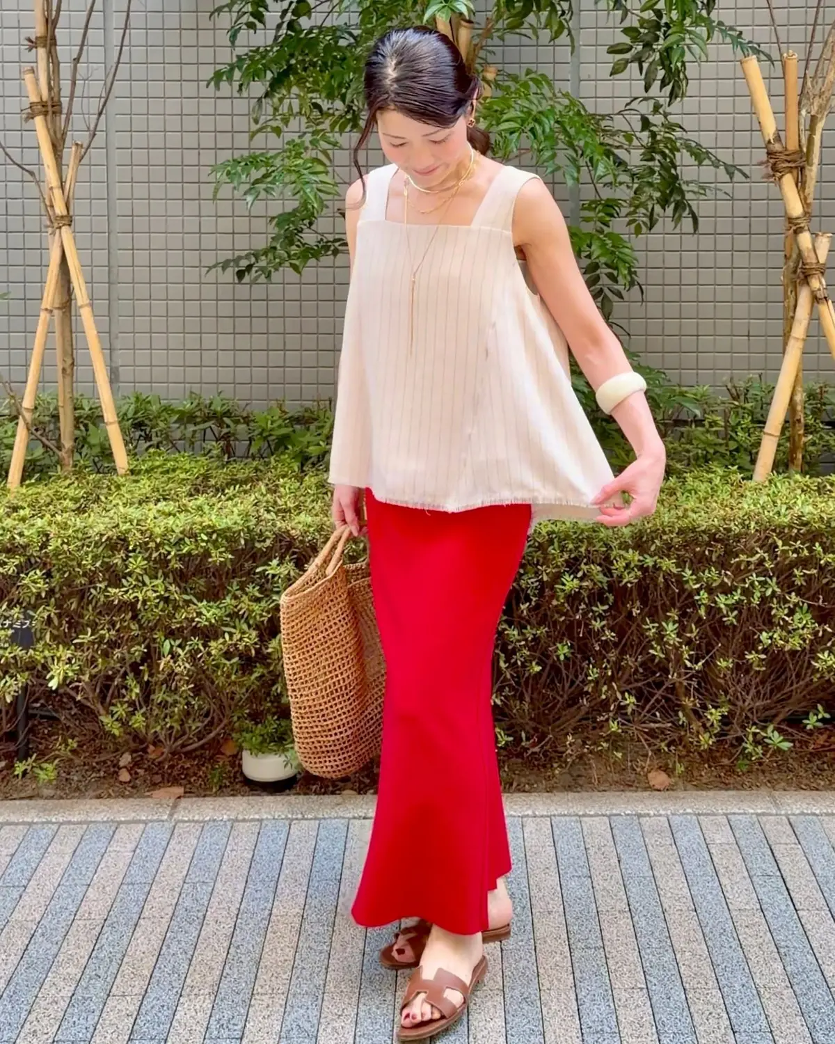 ベージュのトップスと赤のフレアスカートを合わせた女性。茶色のサンダルを履き、編み込みのバッグを持つ。背景には緑の植栽とグレーの壁がある。