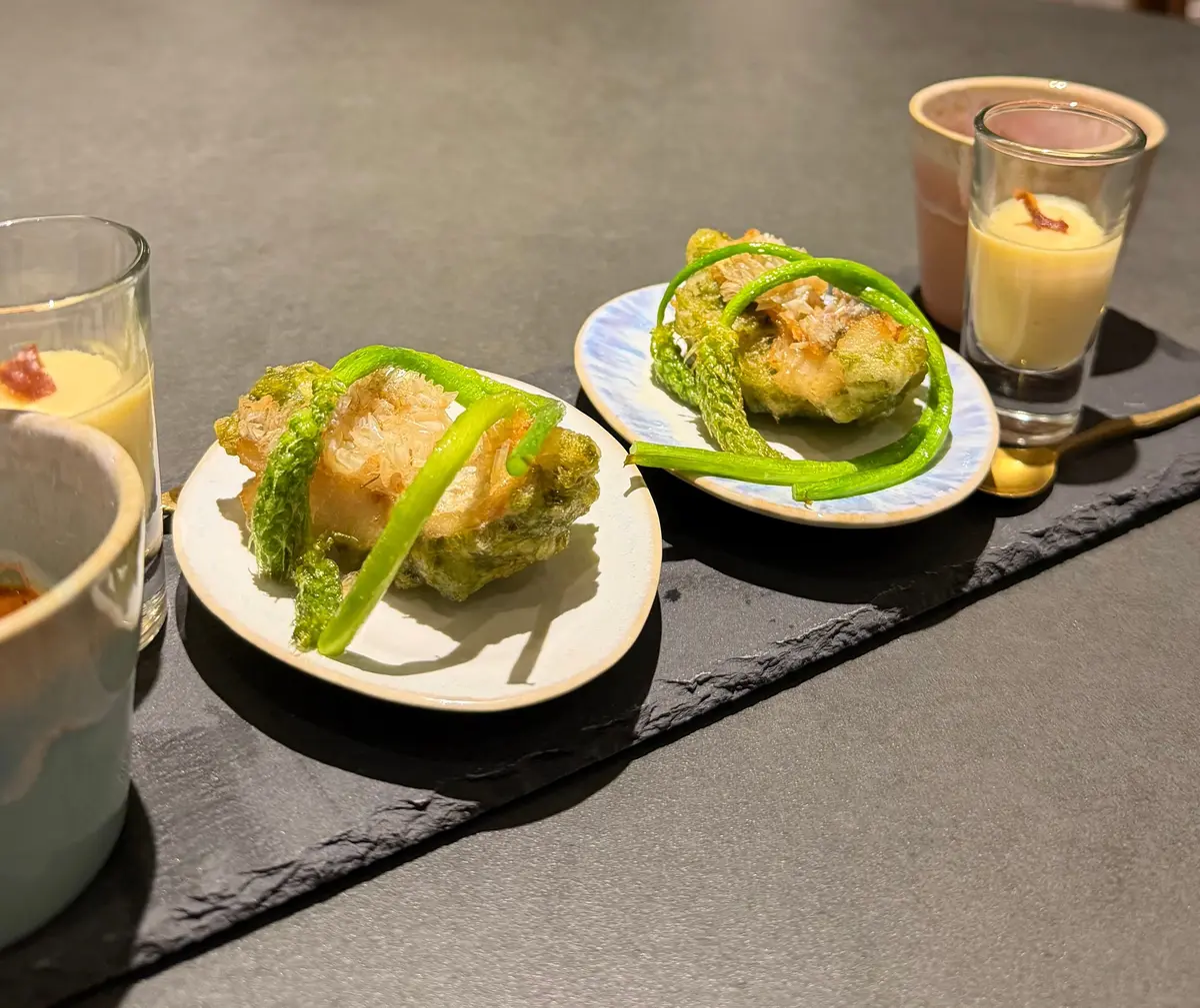 2枚の白い小皿に盛り付けられた、緑色の野菜を添えた揚げ物料理。ガラスのグラスに入ったベージュ色の液体と、そばに置かれた茶色のマグカップも写っている。黒いスレートのトレイに置かれ、暗めのテーブルに配置されている。